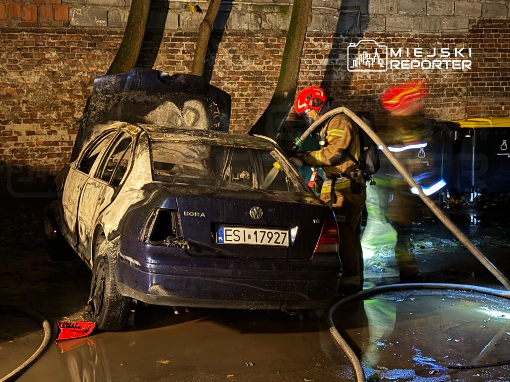 Strażacy gaszą palący się samochód osobowy na miejskim podwórku, w tle ceglana ściana i kontenery na śmieci.