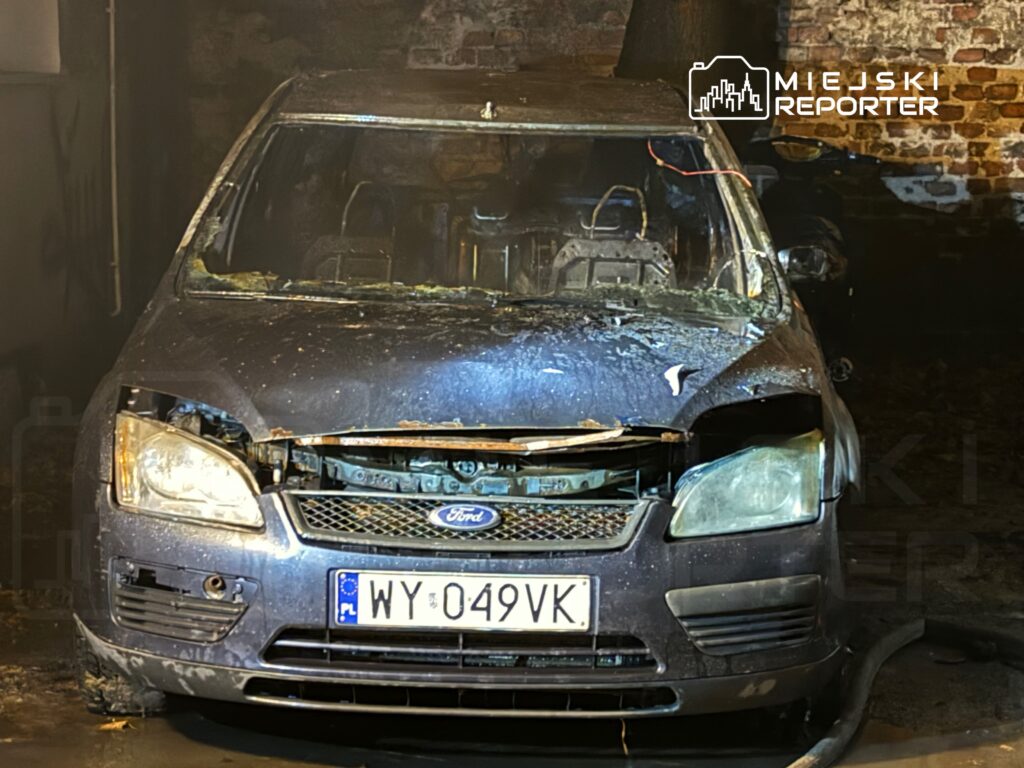 Spalony samochód osobowy marki Ford stoi w ciemnym pomieszczeniu, z widocznymi uszkodzeniami przodu i wnętrza.