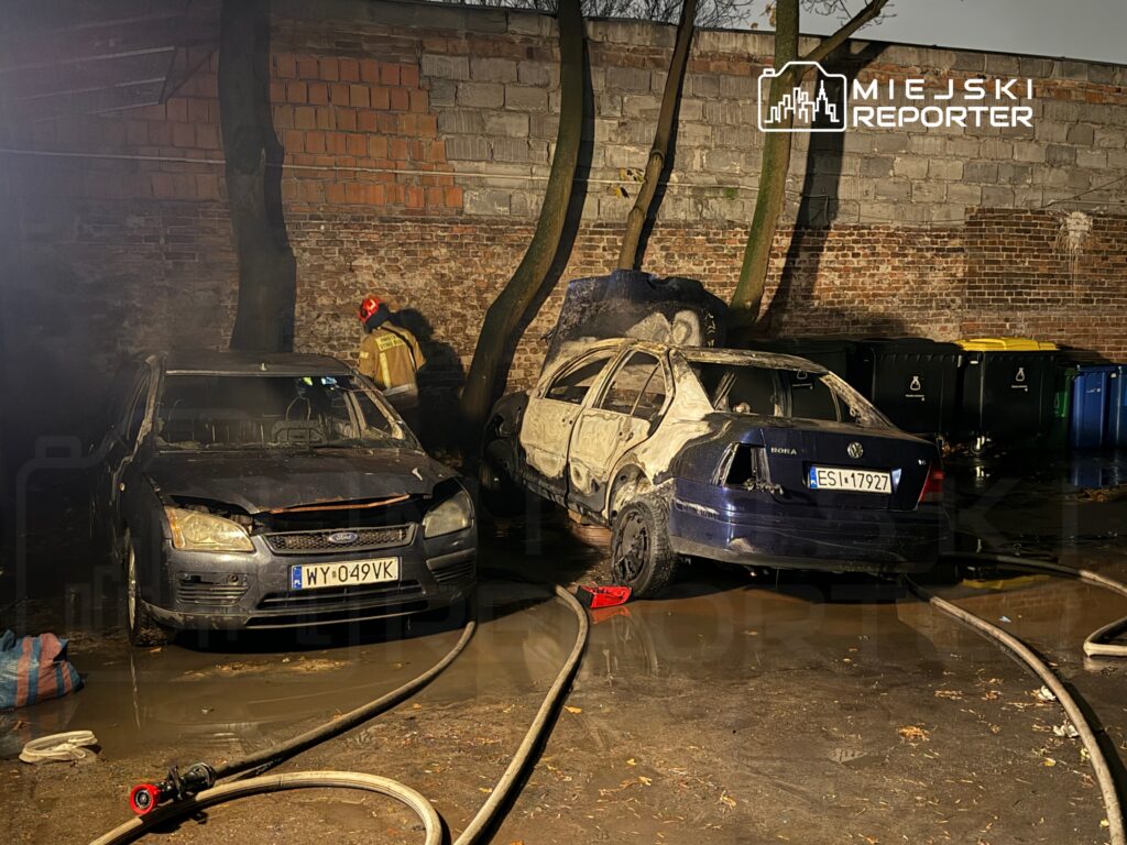 Strażak w hełmie przeszukuje teren przy dwóch uszkodzonych samochodach, w tle widoczne kontenery na odpady.