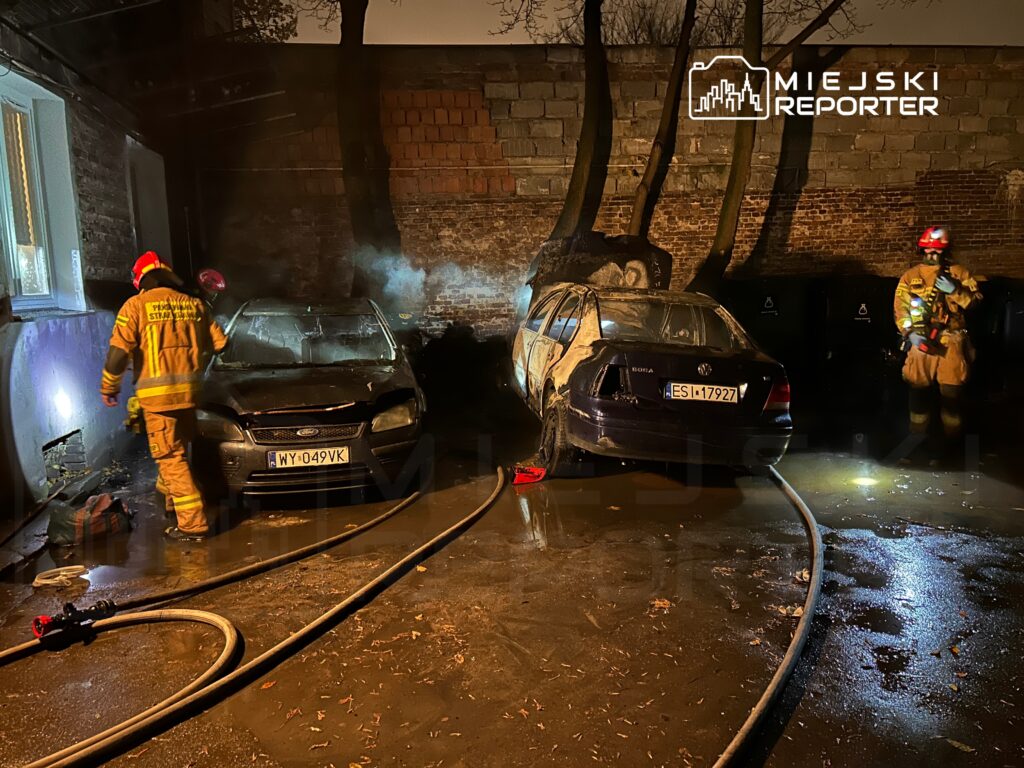 Strażak w uniformie gaśniczym przeszukuje miejsce pożaru, obok dwóch uszkodzonych samochodów w ciemnym otoczeniu.