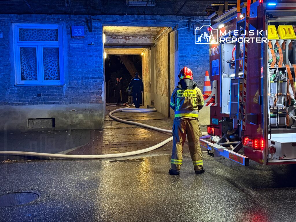 Strażak w hełmie i mundurze obserwuje akcję ratunkową przy wjeździe do podwórza, z wężem strażackim na ziemi.