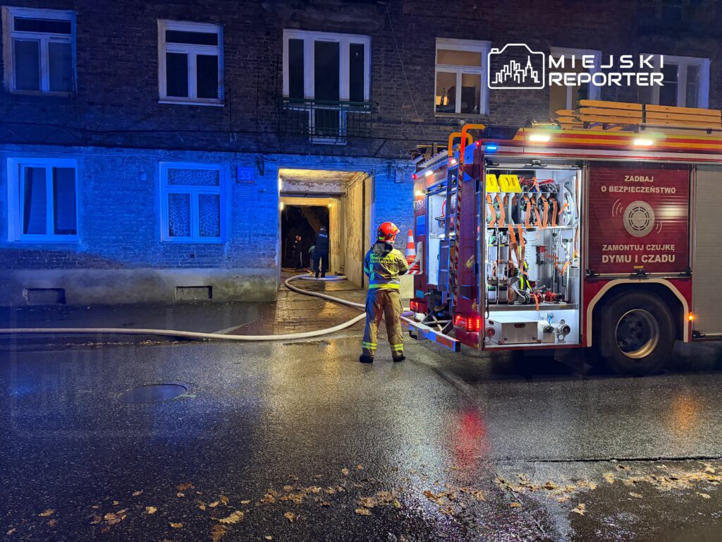 Strażak w hełmie obsługuje wóz strażacki na oświetlonej niebieskim światłem ulicy, obok wejścia do budynku.