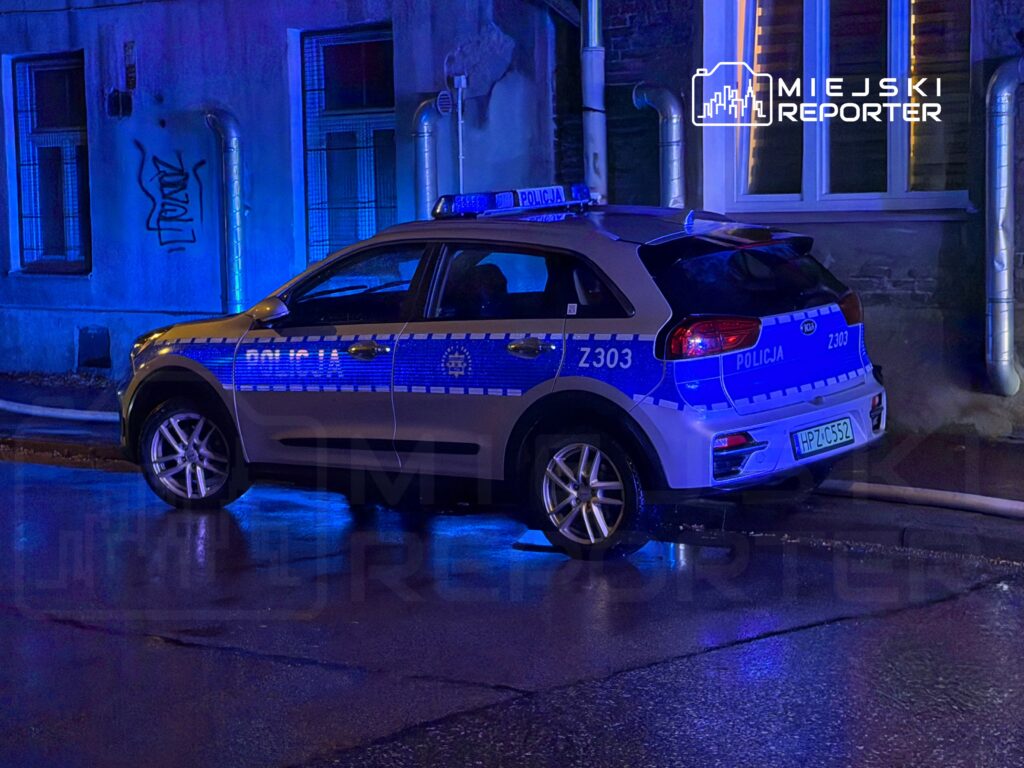 Radiowóz policyjny zaparkowany na mokrej nawierzchni w nocy, oświetlony niebieskimi światłami, obok budynku z graffiti.