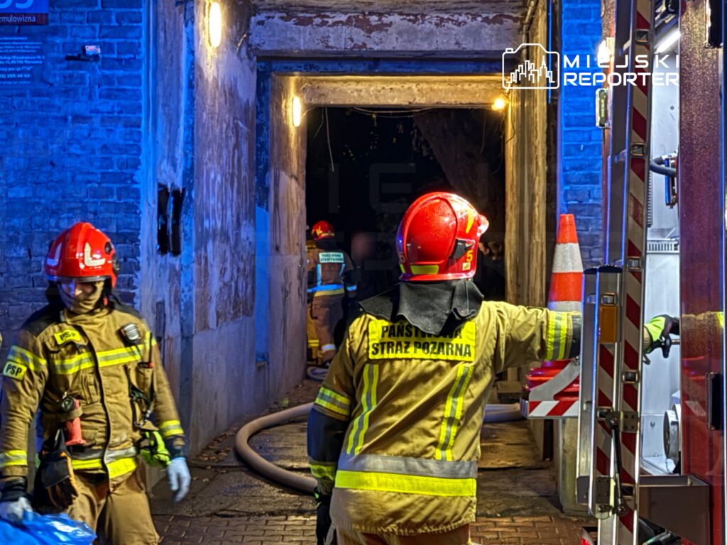 Grupa strażaków w hełmach i mundurach pracuje w ciemnym korytarzu, przygotowując sprzęt do akcji ratunkowej.