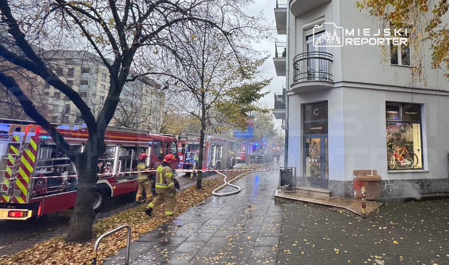 Strażacy gaszą pożar przy budynku mieszkalnym, obok stoją wozy strażackie i wąż gaśniczy na chodniku.