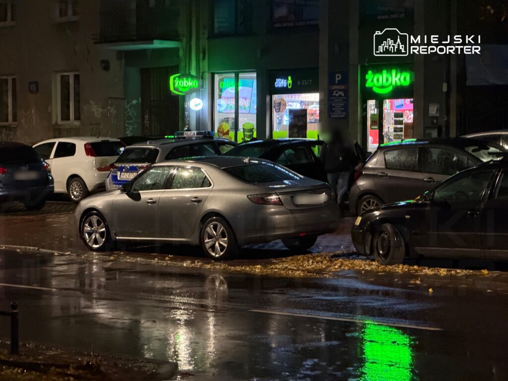 Srebrny samochód zaparkowany na mokrej ulicy, obok sklepu Żabka z podświetlonym znakiem. W tle inne samochody i policja.