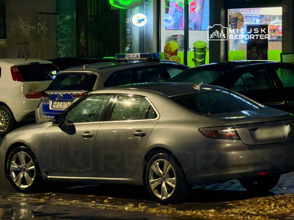 Srebrny sedan zaparkowany na ulicy nocą, w tle widoczny radiowóz policyjny oraz sklep.