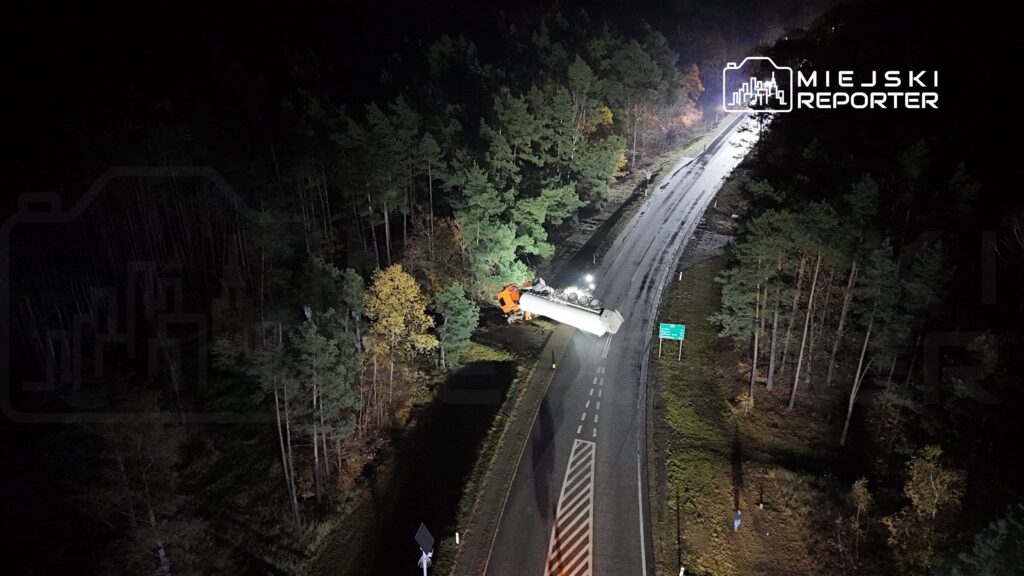 Ciężarówka przewrócona na poboczu drogi, w otoczeniu lasu, oświetlona przez reflektory.