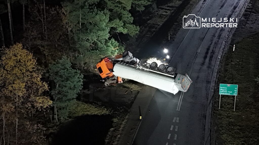 Czerwony ciągnik siodłowy leży na boku obok drogi, a w tle widać oświetlenie i znaki drogowe.
