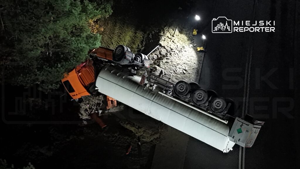 Ciągnik siodłowy przewrócony na poboczu drogi, otoczony przez drzewa, w nocy oświetlony przez latarnie.