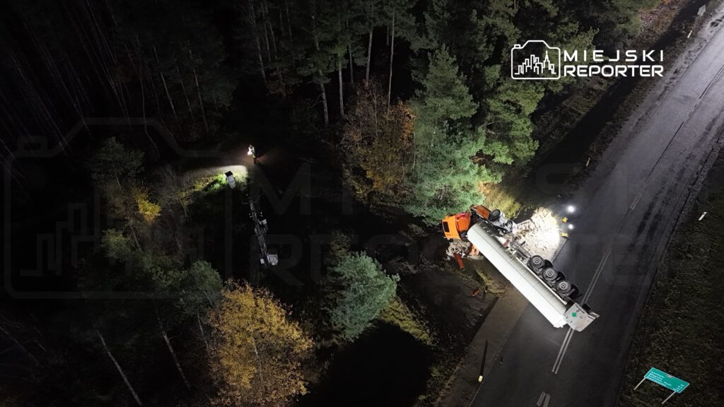 Samochód ciężarowy przewrócony na boku przy drodze, obok pracujący mężczyzna z latarką w lesie.