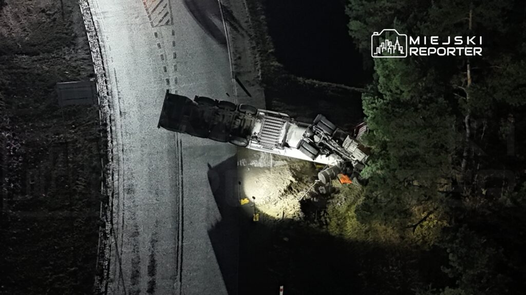 Ciężarówka leży na boku przy drodze, oświetlona przez latarkę, otoczona przez drzewa.