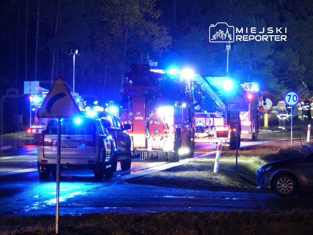 Grupa pojazdów ratunkowych z włączonymi sygnałami świetlnymi na drodze, w tle drzewa i oznakowanie drogowe.