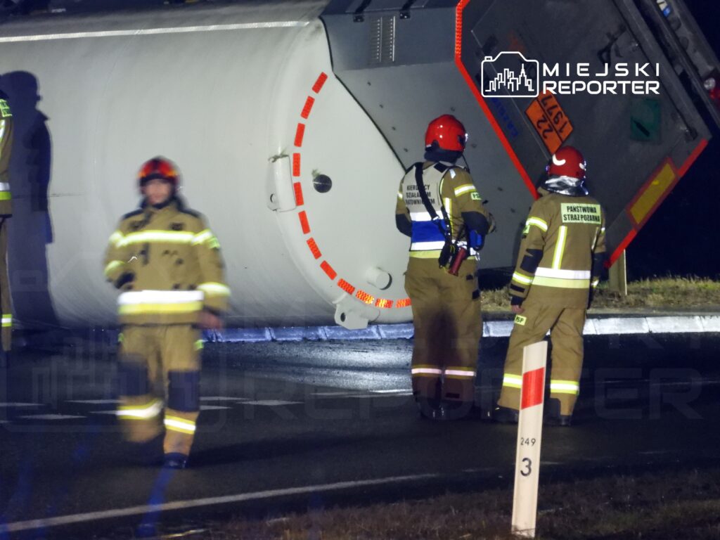 Grupa strażaków w mundurach z odblaskowymi elementami ocenia przewrócony na bok kontener na drodze.