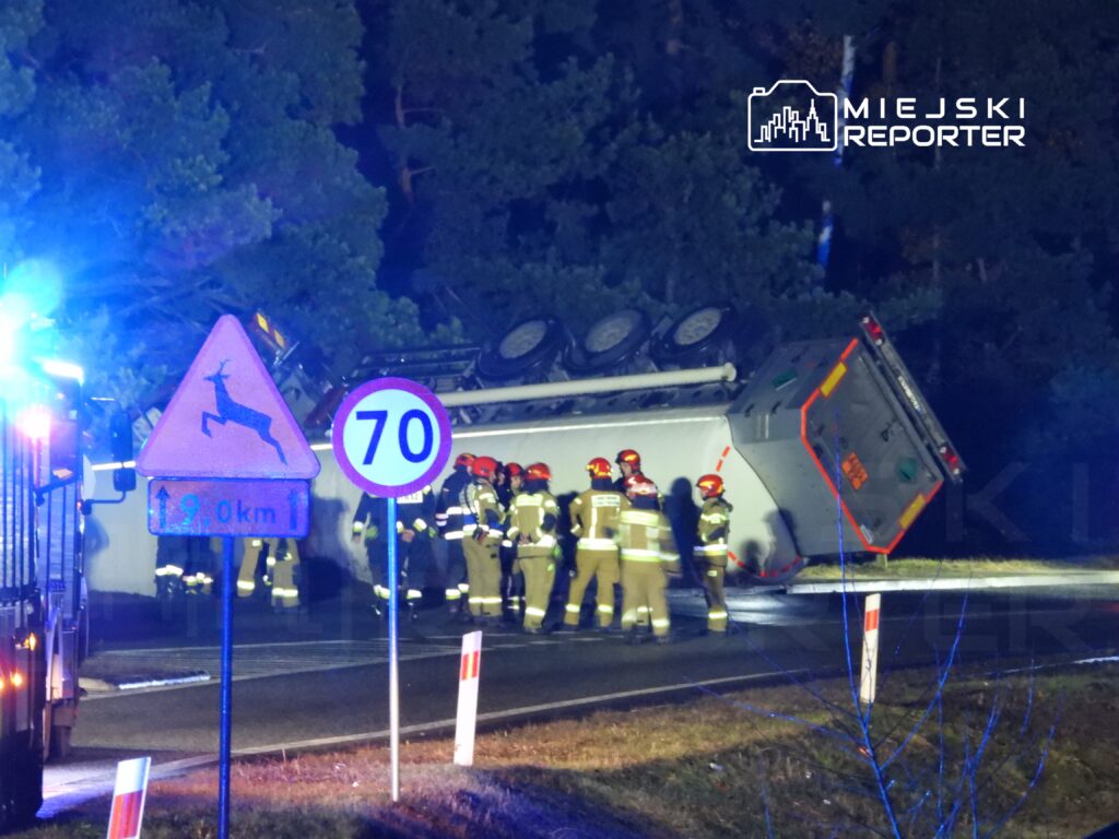 Grupa strażaków w mundurach próbuje pomóc przy przewróconym pojeździe na drodze w lesie, w nocy.