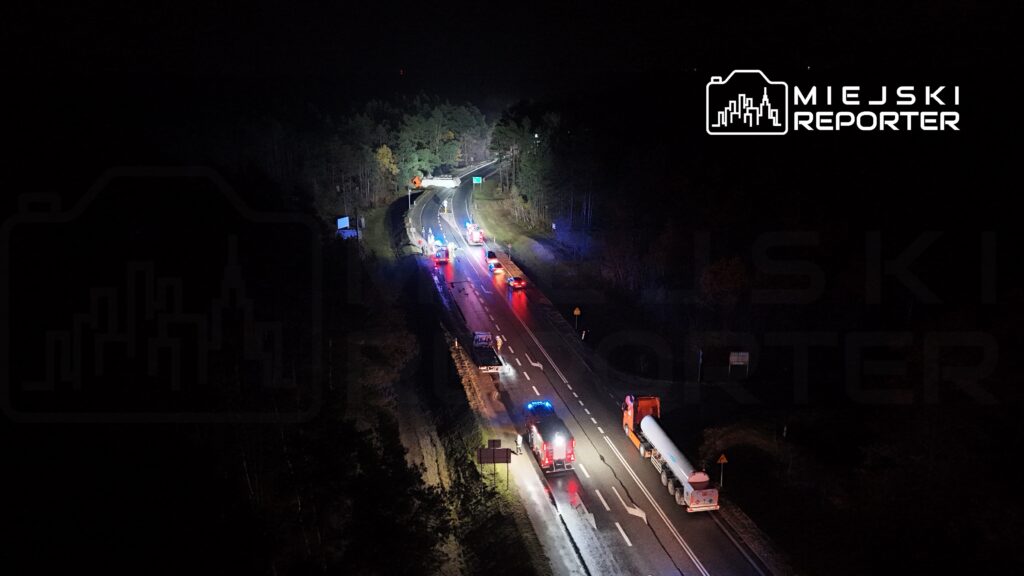 Na drodze nocą widoczne są pojazdy służb ratunkowych oraz policyjnych, a także samochody osobowe z włączonymi światłami.