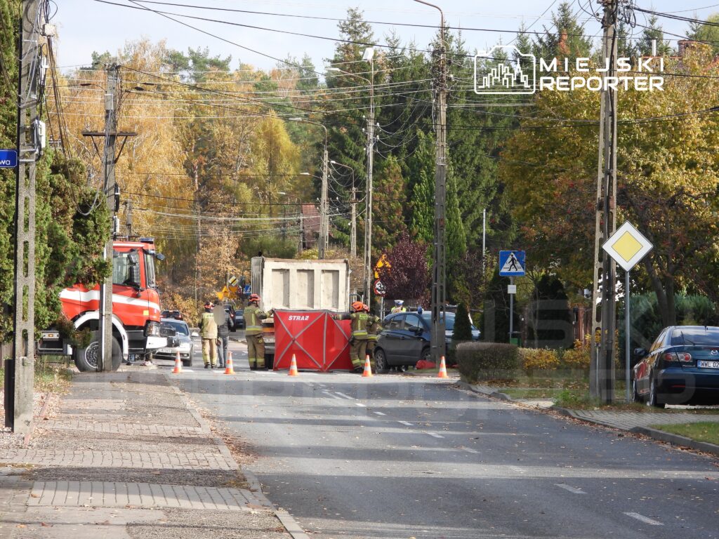 Strażacy pracują przy zablokowanej drodze, obok ciężarówki i kontenera, otoczeni pomarańczowymi pachołkami.