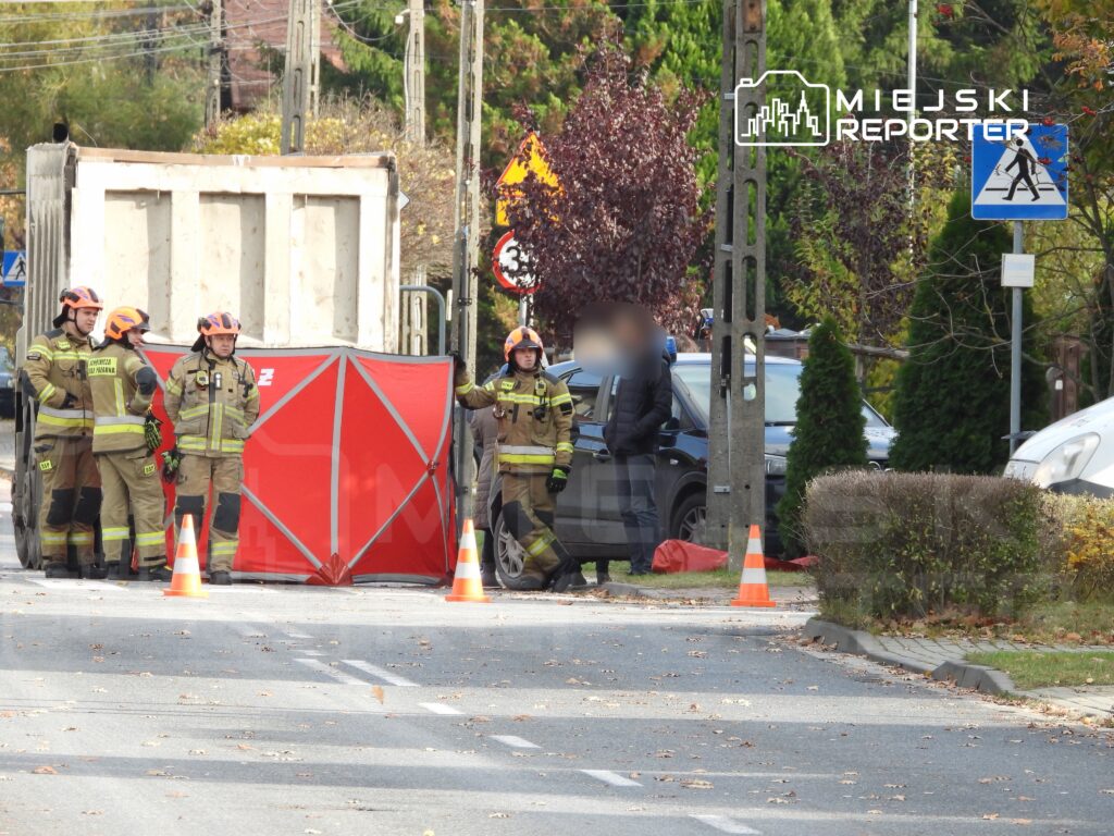 Grupa strażaków w hełmach i mundurach ustawia czerwony parawan na jezdni, zabezpieczając miejsce zdarzenia.