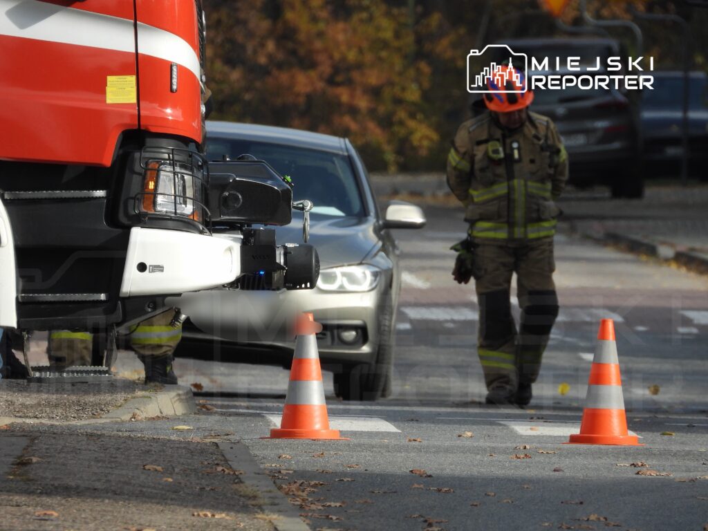 Strażak w mundurze stoi obok wozu strażackiego na ulicy, monitorując sytuację przy oznakowanych pachołkach.