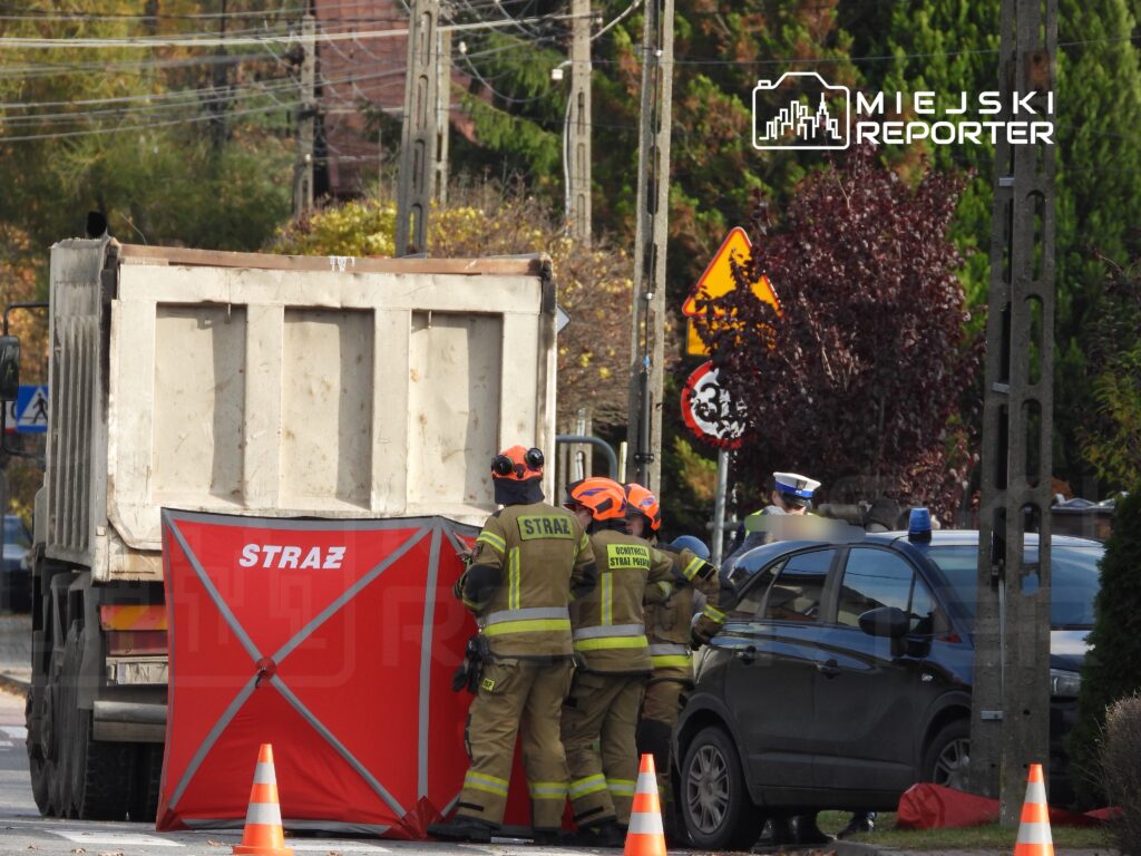 Strażacy w mundurach pracują przy czerwonym namiocie na ulicy, obok zaparkowanego radiowozu.