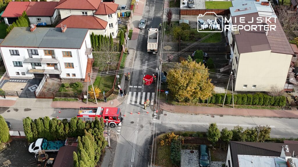 Strażacy pracują na skrzyżowaniu, obok czerwonego wozu strażackiego, zabezpieczając miejsce zdarzenia.