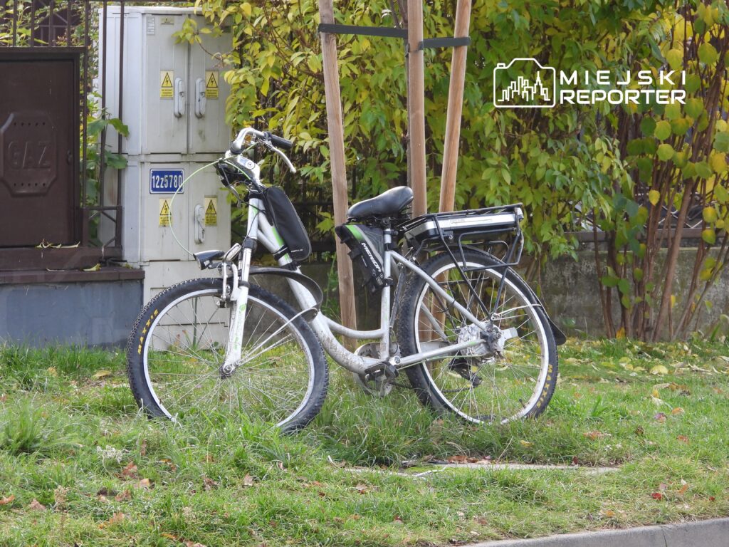 Rower elektryczny zaparkowany na trawie obok metalowej skrzynki z oznaczeniami ostrzegawczymi.