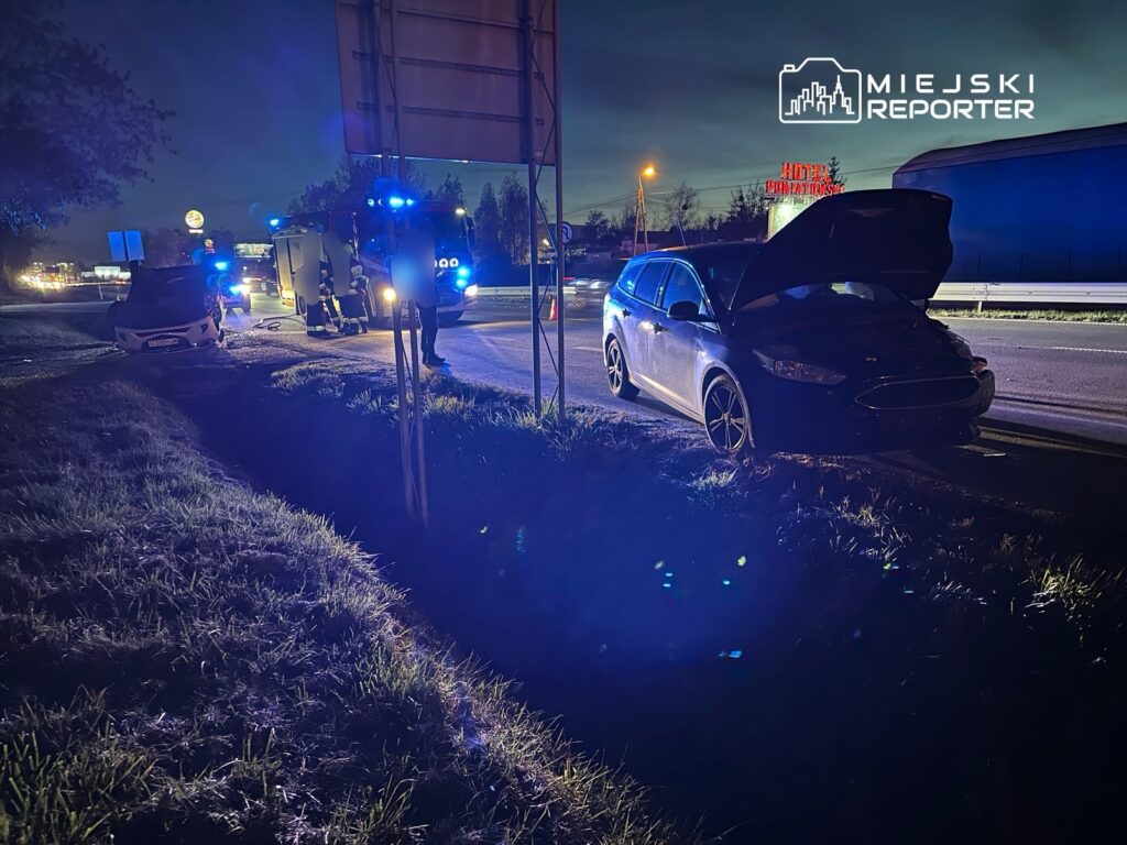 Na drodze stoi czarny samochód z otwartą maską, obok niego zespół ratunkowy z niebieskimi sygnalizatorami.