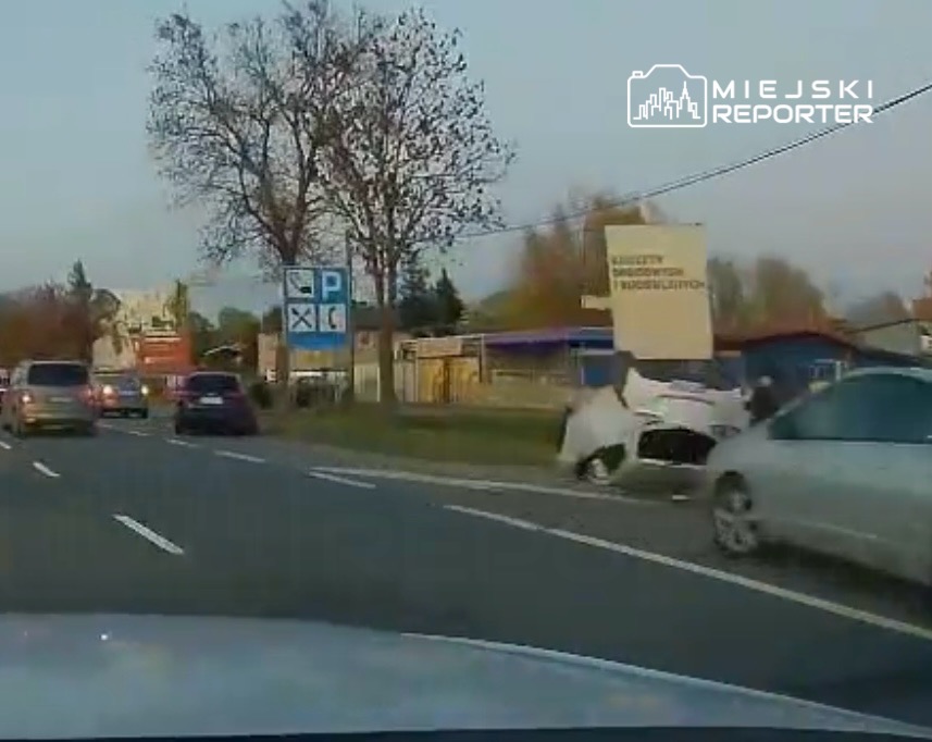 Samochód osobowy leży na boku przy ruchliwej drodze, obok niego stoją mężczyźni, a w tle widoczne znaki drogowe.