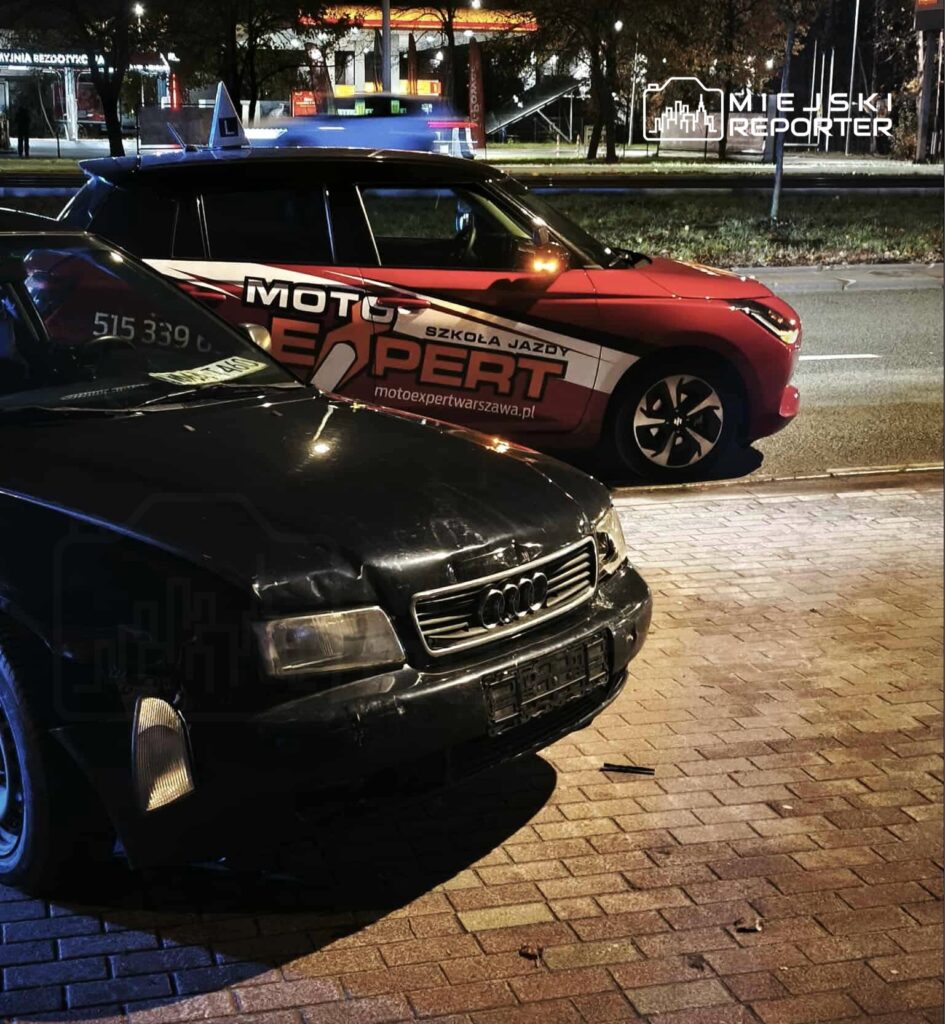 Czarny samochód osobowy marki Audi zaparkowany na chodniku obok czerwonego pojazdu z oznaczeniem szkoły jazdy.