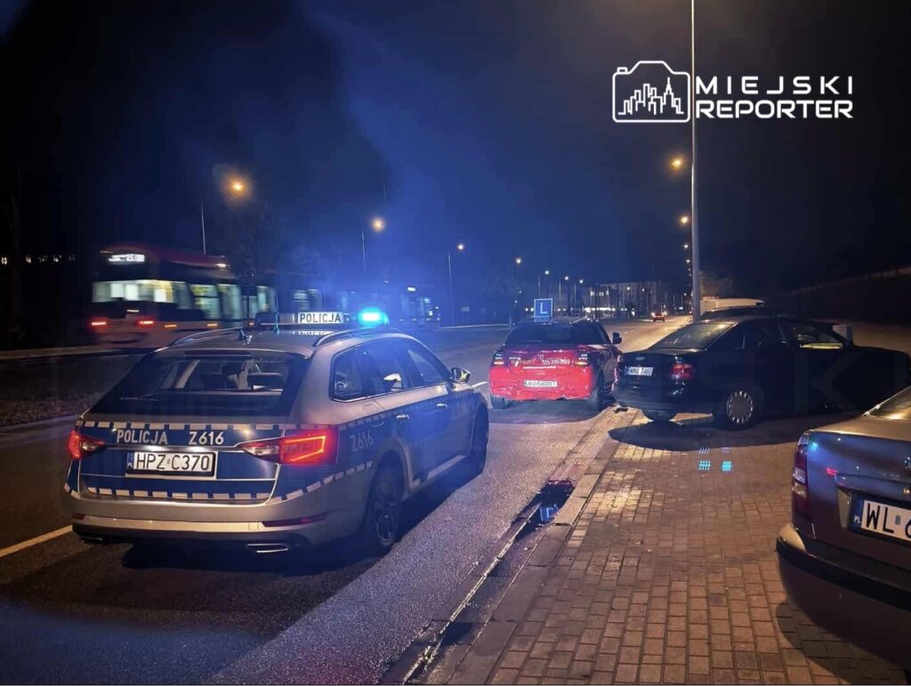 Policyjny radiowóz z włączonymi sygnałami świetlnymi stoi obok dwóch uszkodzonych samochodów na nocnej ulicy.