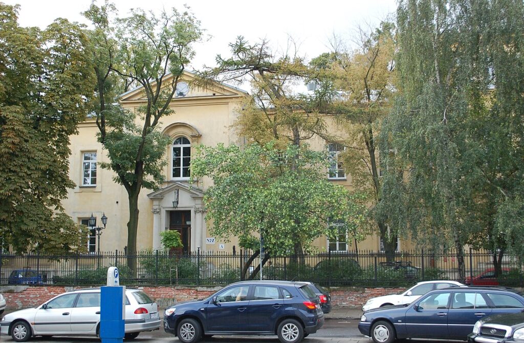 Budynek z jasną elewacją otoczony drzewami, z widocznymi samochodami zaparkowanymi na ulicy.