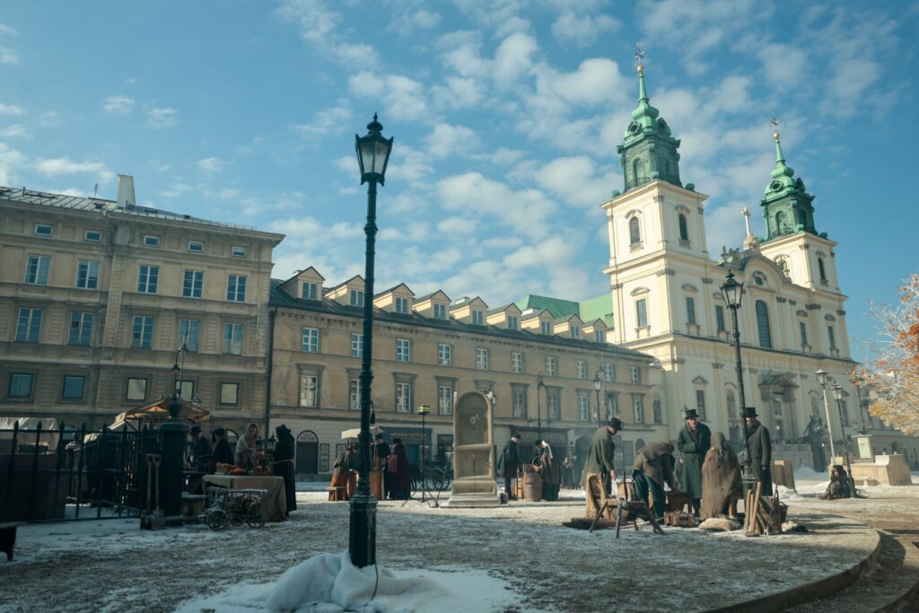 Grupa mężczyzn w historycznych strojach pracuje na ośnieżonym placu, w tle widać barokowy kościół z zielonymi wieżami.