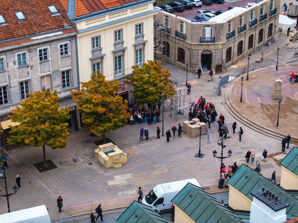 Ludzie gromadzą się na placu w otoczeniu drzew z żółtymi liśćmi oraz budynków, w tle widać pojazdy i rusztowania.