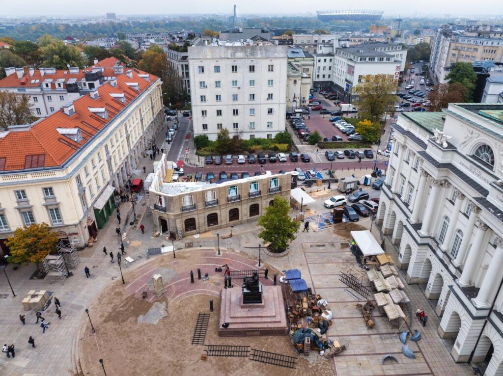 Widok na plac z pomnikiem, w tle nowoczesne budynki i zieleń, w centrum trwają prace budowlane.
