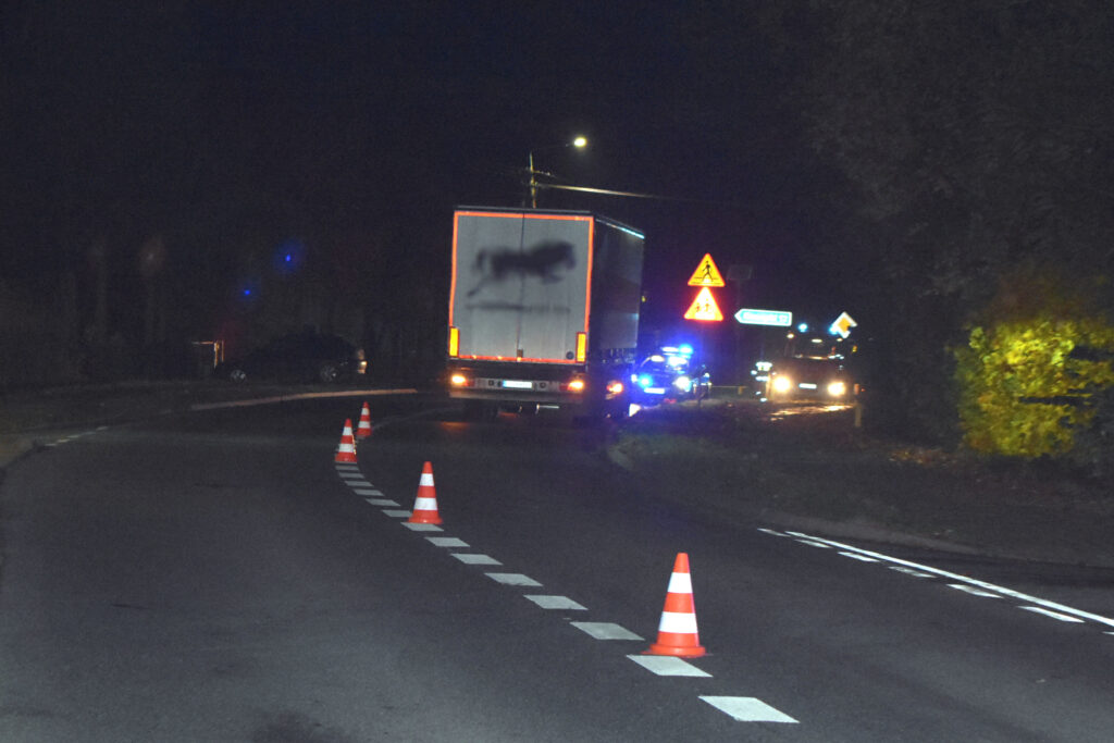 Ciężarówka z oświetleniem awaryjnym zatrzymana na zakręcie drogi, wokół ustawione pachołki drogowe.