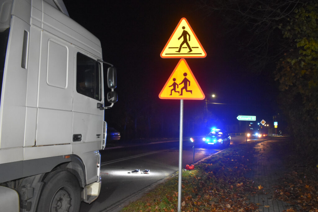Ciężarówka stojąca obok znaków ostrzegawczych dla pieszych i dzieci, w tle policyjne światła na drodze.