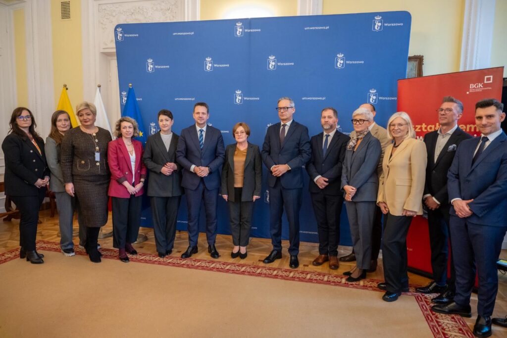 Grupa osób w formalnych strojach stoi w sali z niebieskim tłem i flagami, pozując do zdjęcia.