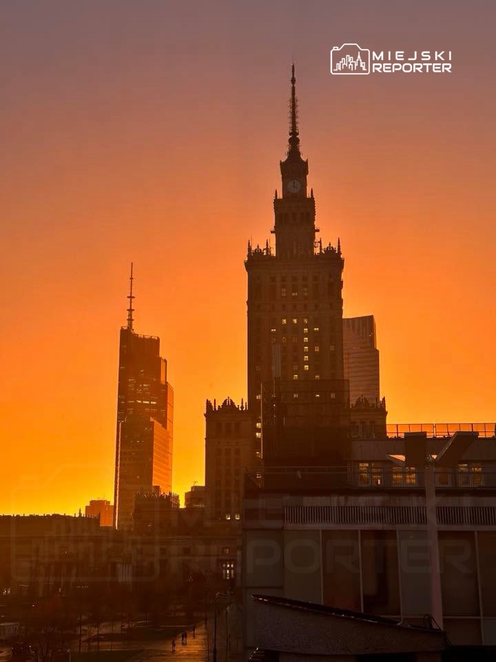 Wysokie budynki w stolicy na tle zachodzącego słońca, tworzące pomarańczową poświatę.