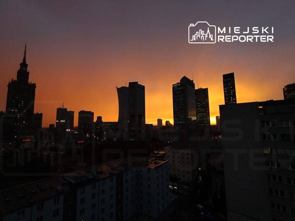 Panoramiczny widok na skyline miasta o zachodzie słońca, z dominującymi wieżowcami i ciepłymi odcieniami nieba.