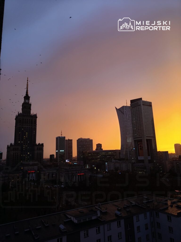 Warszawski krajobraz z dominującą wieżą z zegarem i nowoczesnymi budynkami, oświetlony ciepłym zachodem słońca.