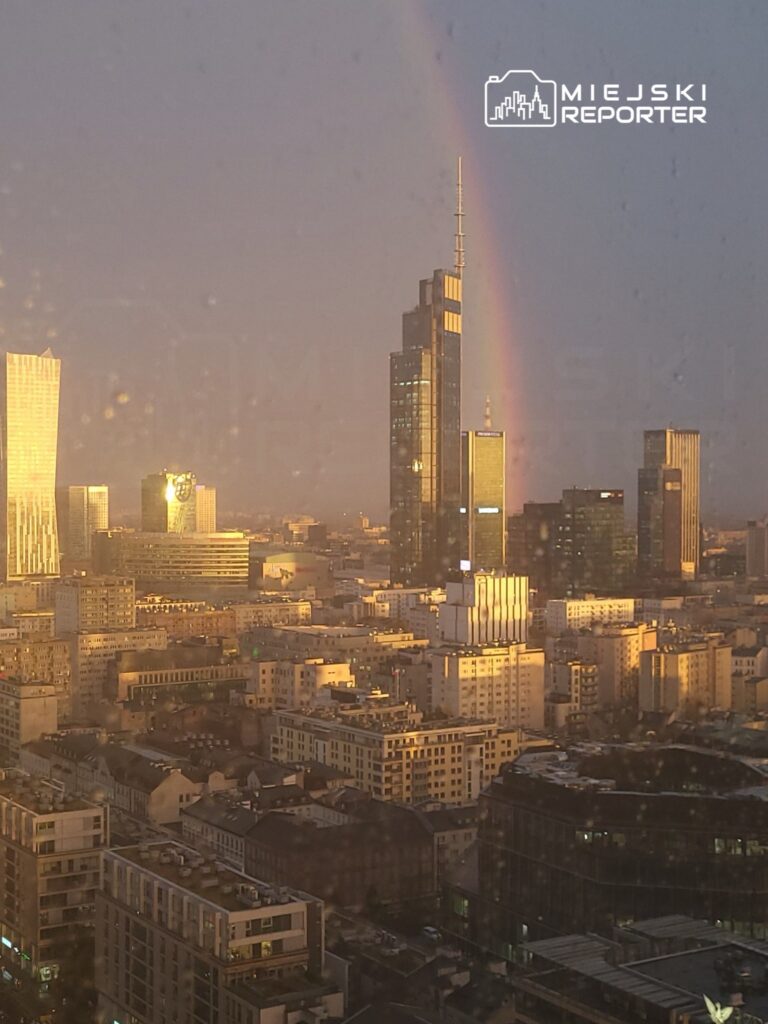 Wysokie wieżowce w miejskim krajobrazie z tęczą na tle zachodzącego słońca.
