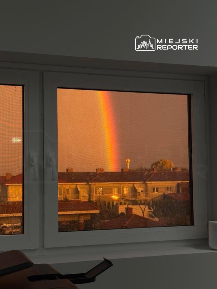 Tęcza pojawiająca się na niebie po deszczu, widoczna przez okno w mieszkaniu, z rzędem domów w tle.