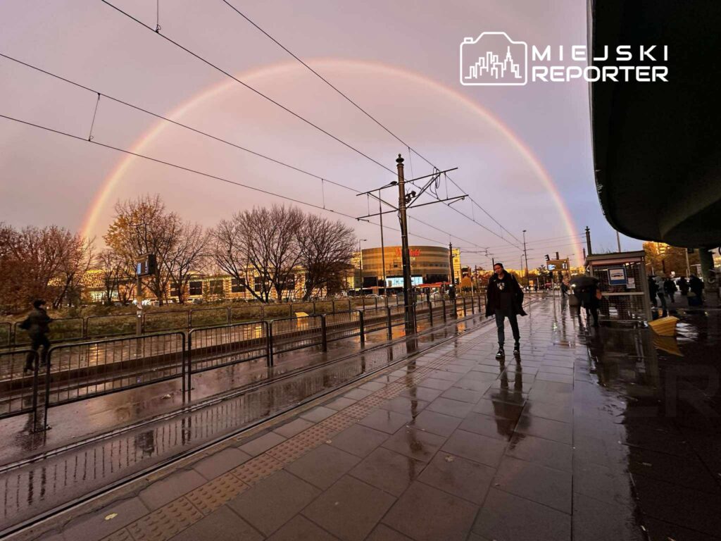 Tęcza rozciągająca się nad miejskim krajobrazem, z ludźmi spacerującymi po mokrej nawierzchni chodnika.