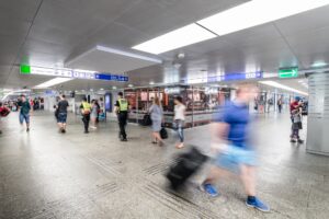 Grupa ludzi poruszająca się w podziemnym korytarzu stacji, z oznaczeniami kierunków i punktami usługowymi w tle.