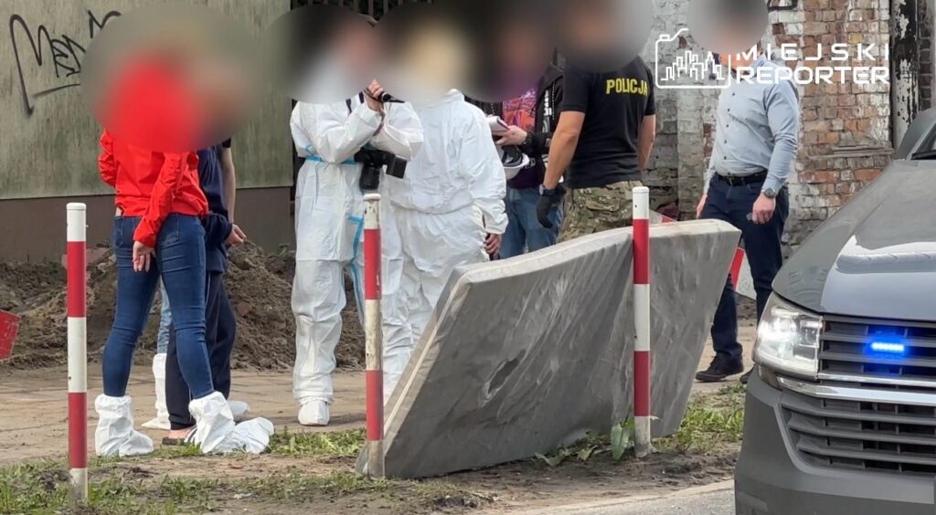 Grupa osób w odzieży ochronnej rozmawia w pobliżu betonowej płyty, w tle widoczny radiowóz i zniszczona ściana budynku.