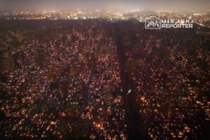 Widok z lotu ptaka na cmentarz, w którym setki zniczy oświetlają groby w ciemności, tworząc świetlną mozaikę.