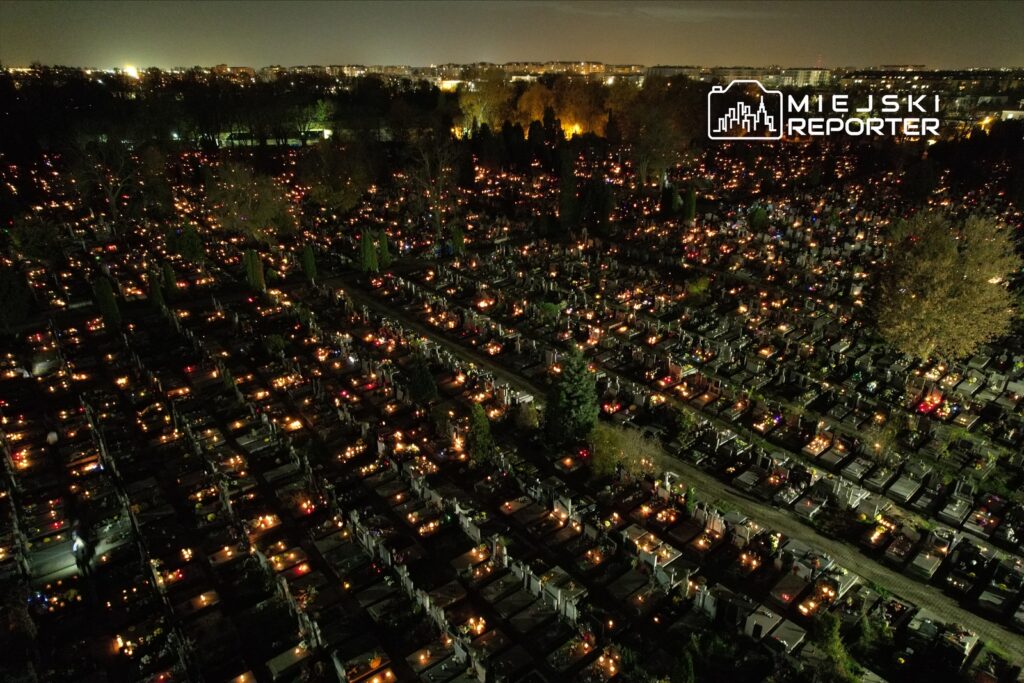 Cmentarz oświetlony setkami zniczy, z widocznymi postaciami odwiedzających groby, w tle nocne niebo i miasto.