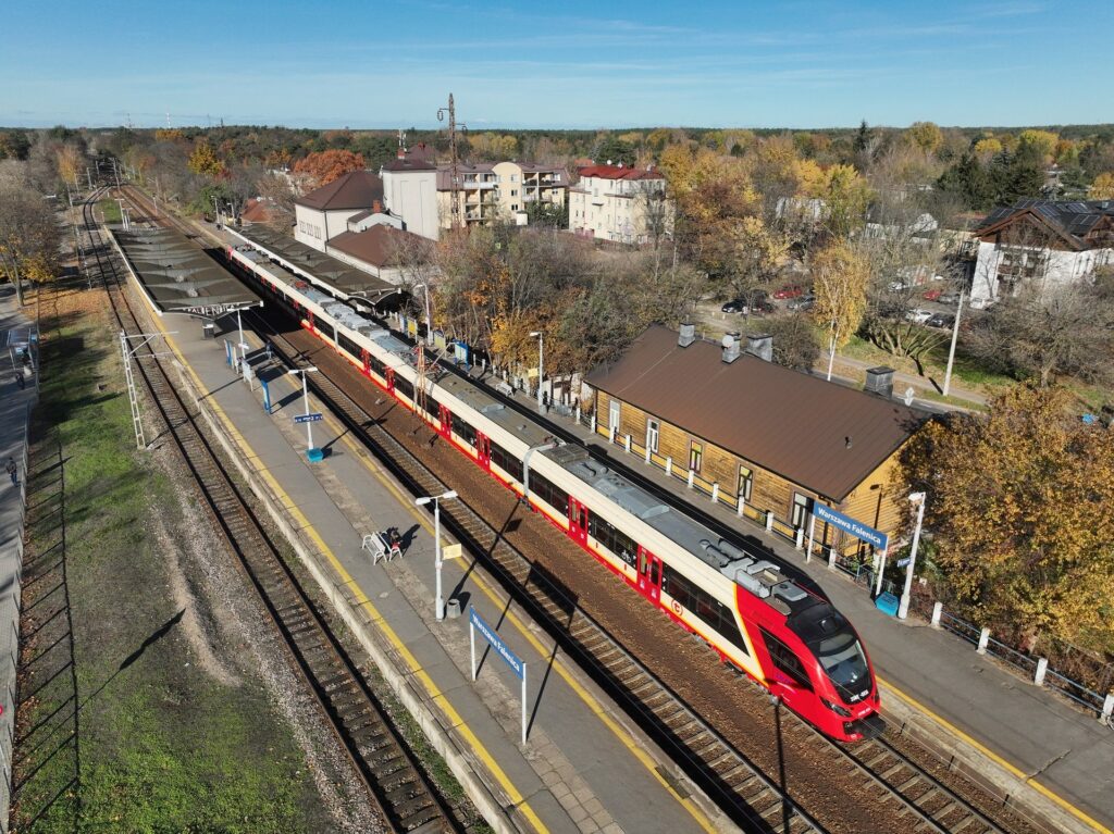 Pociąg osobowy w czerwono-białych barwach zatrzymany na stacji, obok peronu stoją pasażerowie i budynki mieszkalne.