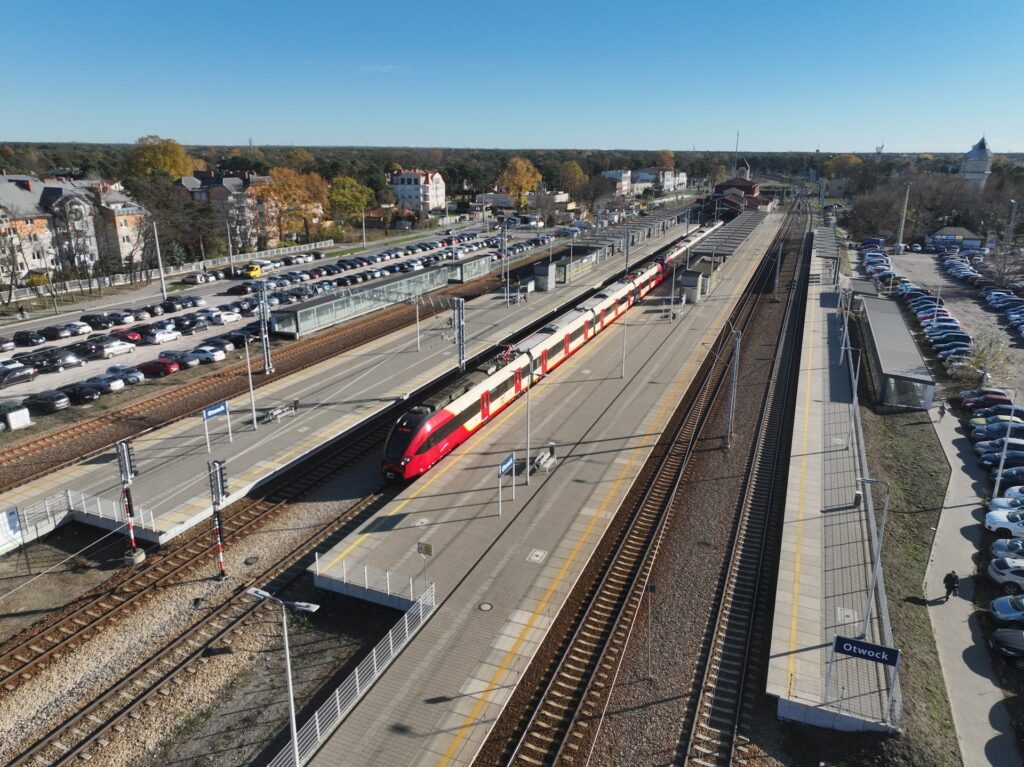 Pociąg w czerwono-żółtych barwach zatrzymuje się na stacji kolejowej, obok parkingu pełnego samochodów.