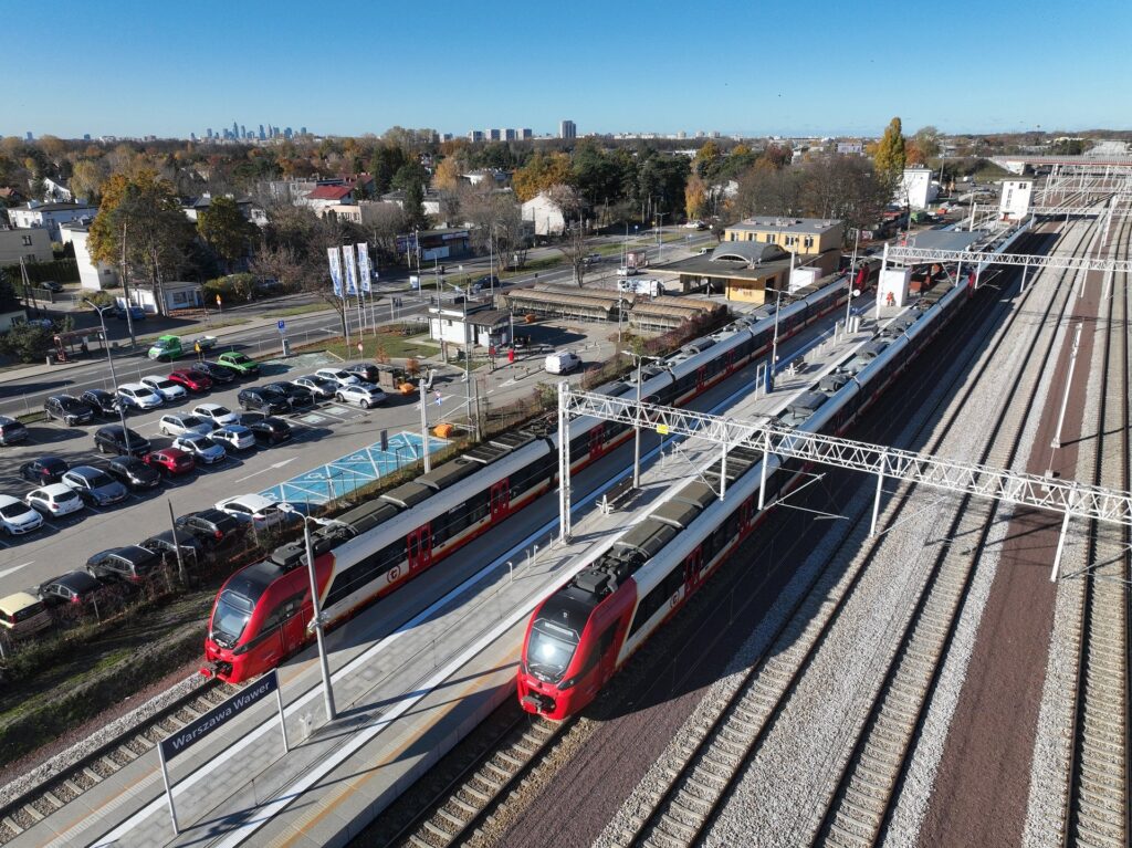 Dwa pociągi na stacji kolejowej, obok zaparkowane samochody oraz widok na miasto w tle.